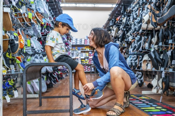 Mother smiling while assisting her son in trying on new shoes at a shoe store, surrounded by a wide selection of footwear, enjoying quality time together during their shopping experience