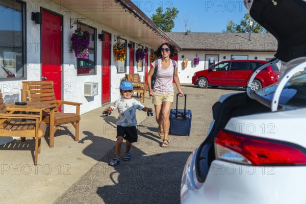 Happy mother pulling a suitcase while her cheerful son runs alongside, playing with toy cars, arriving at a motel during a fun filled summer road trip vacation