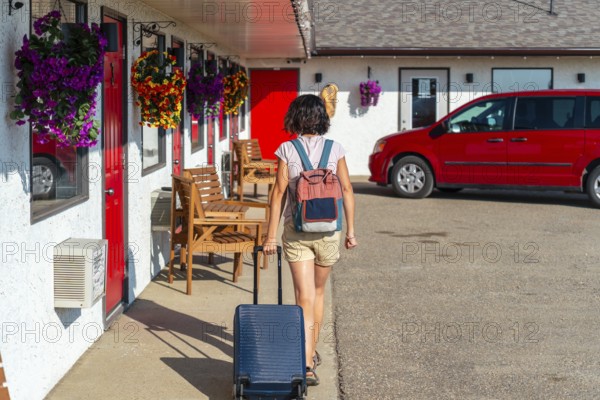Female tourist walking towards her room at a roadside motel, pulling rolling luggage and wearing a backpack under the warm sun, enjoying the journey on a beautiful summer day