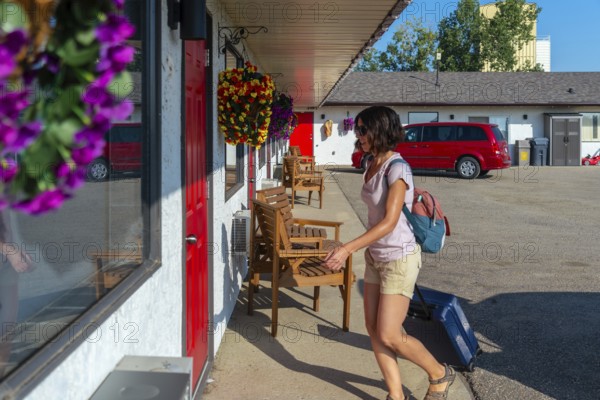 Female tourist is walking to her motel room pulling her wheeled suitcase, carrying a backpack and passing by some wooden chairs on a sunny summer day