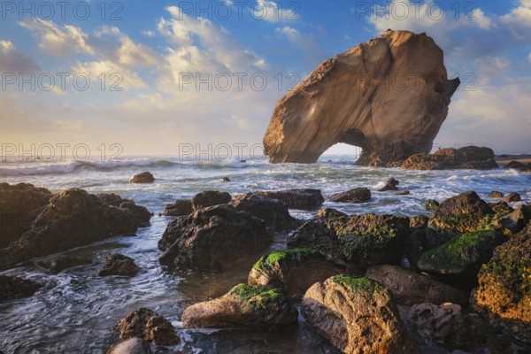 Penedo do Guincho, a large boulder rock arch at Praia da Santa Cruz, Portugal, with ocean waves and sandy beach on sunset