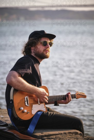 Hipster street musician in black playing electric guitar in the street sitting on pier embankment on sunset