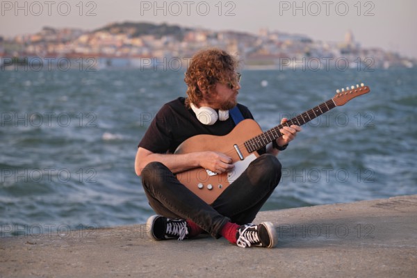Hipster street musician in black playing electric guitar in the street on sunset on embankment with Lisbon in background. Portugal