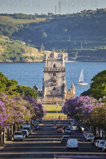 Scenic view of Belem Tower in Lisbon, Portugal, seen over a street with blooming purple jacaranda flower trees street with tourist sailboat on the Tagus River. Portugal
