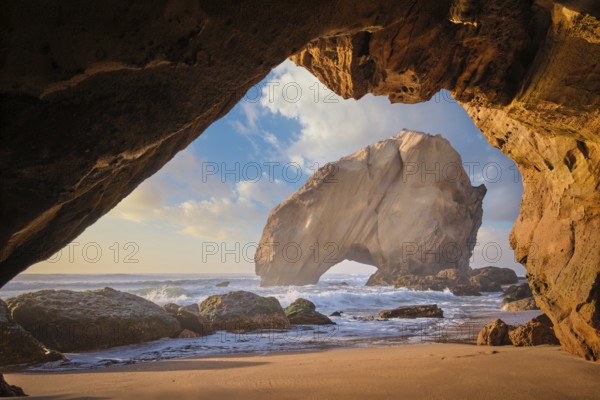 Penedo do Guincho, a large boulder rock arch at Praia da Santa Cruz, Portugal, with ocean waves and sandy beach as seen from grotto cave on sunset