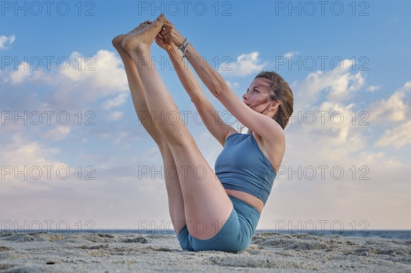 A woman in Ubhaya Padangusthasana, or Double Toe Hold, balancing gracefully on the sandy beach. The sunset sky creates a serene backdrop, highlighting her flexibility and concentration
