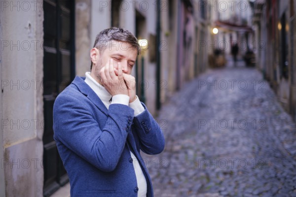 Man musician in a blue blazer and white sweater playing blues on a harmonica with eyes closed, standing outdoors in city street