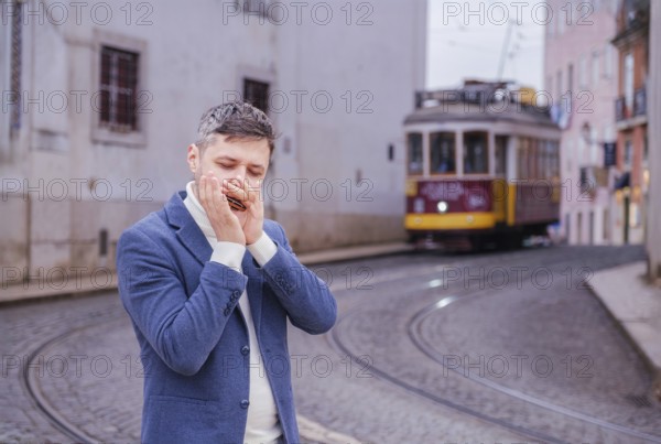 Man musician in a blue blazer and white sweater playing blues on a harmonica with eyes closed, standing outdoors in Lisbon city street, Portugal with tram in background