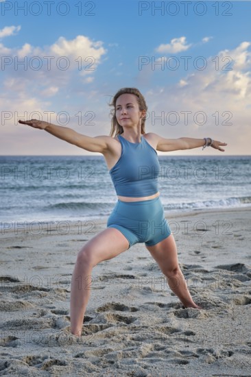 A woman performs Virabhadrasana 2 Warrior 2 pose on the beach, embracing the calm sunset and ocean waves in Ashtanga Vinyasa Yoga