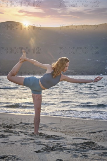 A woman performs Natarajasana, or Dancer's Pose, on a beach during sunset. The sun sets behind mountains, creating a beautiful, peaceful atmosphere as she balances on one leg
