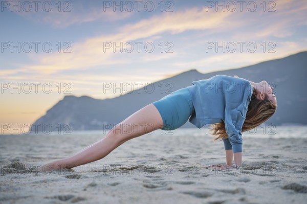 A woman practices Purvottanasana, or upward plank pose, on the beach during sunset. The golden sky reflects on her focused core work as she balances on the sand in serene surroundings