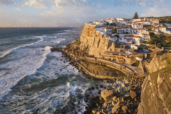 Charming Azenhas do Mar, a picturesque fishing village perched on cliffs. The sun sets over the Atlantic, casting warm hues on the whitewashed buildings and rocky coastline. Portugal