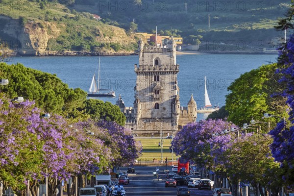 Scenic view of Belem Tower surrounded by blooming jacaranda trees, with sailboats on the Tagus River during sunset in Lisbon, Portugal