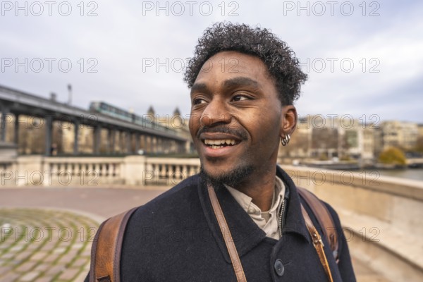 Happy black man smiling and looking away while strolling across paris bir hakeim bridge with a metro train passing, enjoying an urban citybreak by the seine