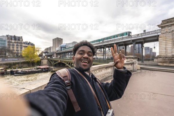 Happy african american man tourist taking a selfie portrait on a bridge over the seine river in paris, france, with the elevated metro passing by in the background