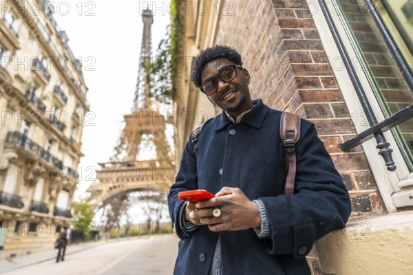 African man enjoying his travel experience in paris, standing on the street with the eiffel tower in the background, smiling while holding and interacting with a mobile phone