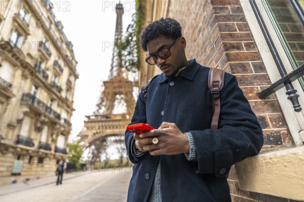 Young black man in paris leaning against a building, focusing on his smartphone, representing travel, technology, and urban modern connectivity in a famous european city