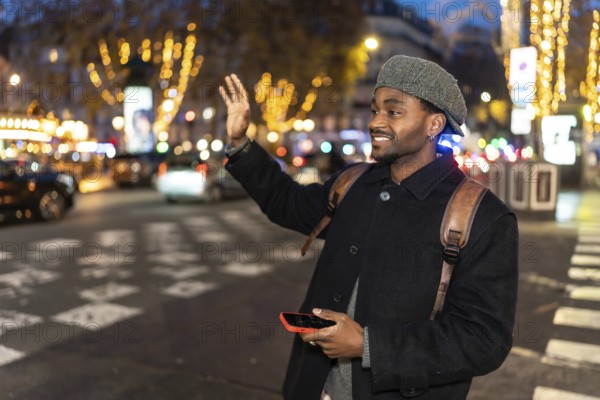Young man smiling while hailing a taxi on a vibrant city street at night, holding a smartphone and wearing a backpack and hat, surrounded by blurred evening lights