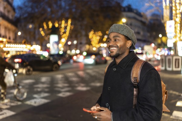 Smiling young black man wearing a winter coat and beret, carrying a backpack, using a phone while walking on a city street at night with blurred lights in the background