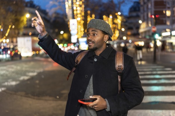 Young man in a winter coat stands on a busy city street at night, holding a smartphone and raising his hand to hail a taxi amid blurred urban lights and traffic