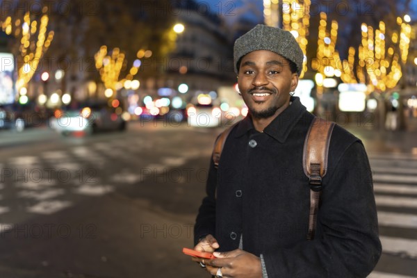 Young black man smiling in an urban setting at night, holding a mobile phone and standing on a city street with blurred car lights and decorated trees in the background