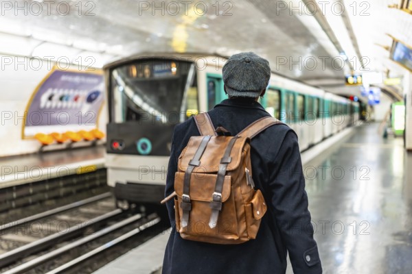 Man with backpack and flat cap standing on a subway platform, watching a train arriving, representing urban travel, commuter lifestyle, and city adventure