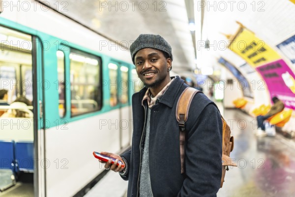 Young black man in a beret smiles at camera while holding a phone and backpack on a paris subway platform, waiting for a train urban commuter enjoying travel and tech convenience