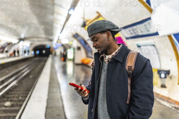 Young black man in a flat cap and backpack checks his smartphone while waiting on a busy urban subway platform, commuting and browsing apps during his daily journey