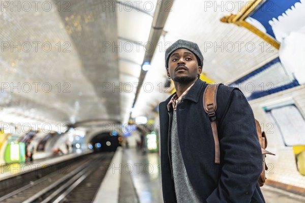 Young black man in a beret with a backpack stands on a tiled paris metro platform, looking up while waiting for a train, thoughtful and ready for urban travel and exploration
