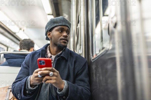 Young black man seated on subway, dressed for winter, holding smartphone and gazing out the window while commuting through the city, thoughtful and connected