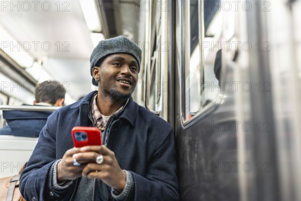 Young black man in beret and jacket rides the subway holding a red smartphone, smiling and looking out the window while commuting through the city, browsing and enjoying the ride