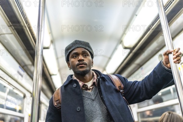 Young black man in winter coat and hat stands holding a subway pole on a crowded metro, looking away pensively during a daily commute, urban travel and routine