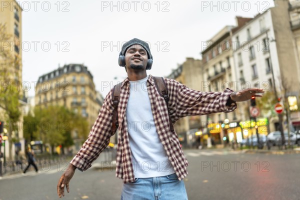 Young black man with headphones and backpack enjoying music and dancing freely with arms outstretched in a plaid shirt on a paris city street, celebrating happiness and urban freedom