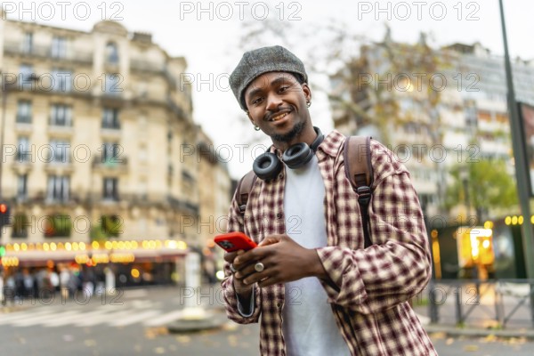 Young african man with a beret and headphones smiling at the camera while checking his smartphone, standing outdoors on a sunny day in the vibrant streets of paris