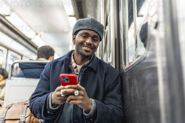 Young black man with a stylish hat smiling and looking at the camera while commuting on a subway train, leaning against the window and holding his mobile phone