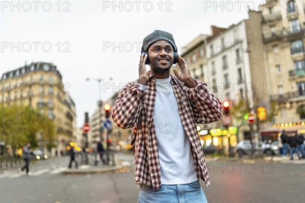 Young handsome black man walking a sunny european city street, smiling while listening to wireless headphones, relaxed and stylish in a plaid shirt, beanie and backpack