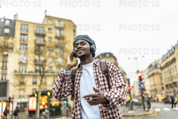 Young african man smiling and listening to music with headphones while walking in a bustling european city street, enjoying a carefree moment of urban life and travel