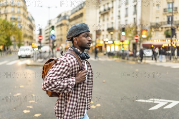 Young black man standing on a city street at a crosswalk, wearing a hat and backpack with headphones around his neck, looking right while waiting to cross