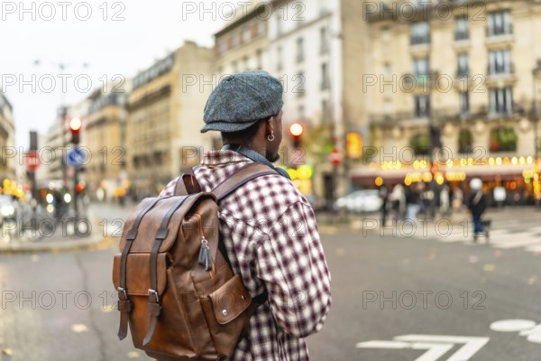 Young man wearing a cap and brown backpack stands on the curb of an urban street, looking across the busy intersection, ready to continue his journey through the city