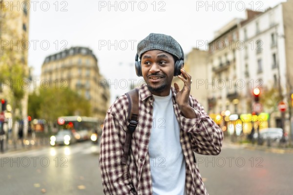 Young black man enjoying music through headphones while walking on an urban street, adjusting his headwear and smiling, with blurred city buildings and vehicles in the background