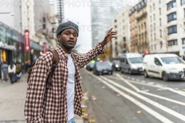 Young black man wearing a beret and a plaid shirt standing on an urban sidewalk, raising his hand to hail a taxi on a busy city street with traffic moving in the background