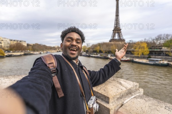 Young male tourist smiling and gesturing with open hand, capturing a memorable selfie while enjoying a vacation on a bridge overlooking the seine river with the iconic eiffel tower