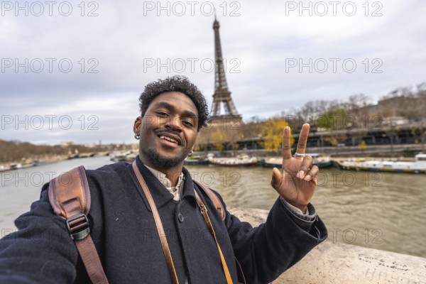 Smiling young man vlogging a selfie on a paris trip, flashing a peace sign with the eiffel tower and seine river in the background, bundled in a coat and backpack
