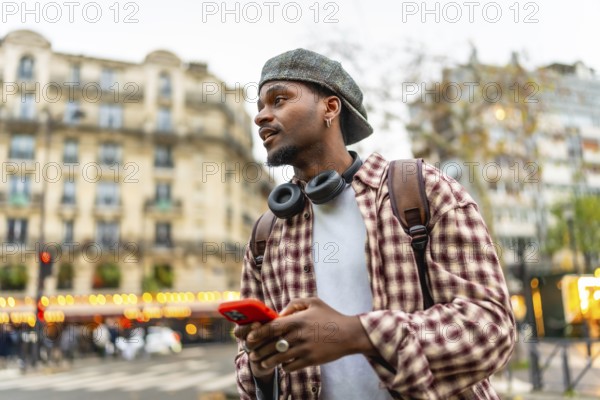 Young man using a smartphone and wearing headphones, a backpack, and a casual plaid shirt while exploring a city street with historic architecture in the background