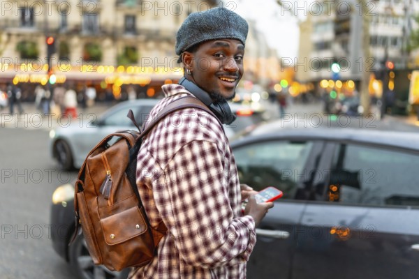 Young black man smiling, wearing a beret and backpack, holding a smartphone while walking on a busy city street with cars and buildings in the background