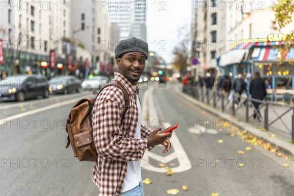 Young black man in a checkered shirt and backpack smiles at the camera while checking his phone on a busy city street, capturing modern urban life and connectivity