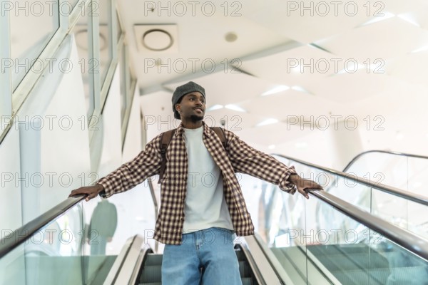 Young black man riding a modern escalator in a downtown shopping mall, wearing a backpack and stylish casual clothes, looking ahead while exploring urban retail spaces