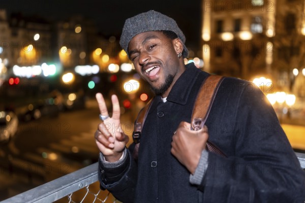 Young man in a flat cap standing outdoors in the city at night, winking at the camera and showing a peace sign, expressing happiness and carefree travel