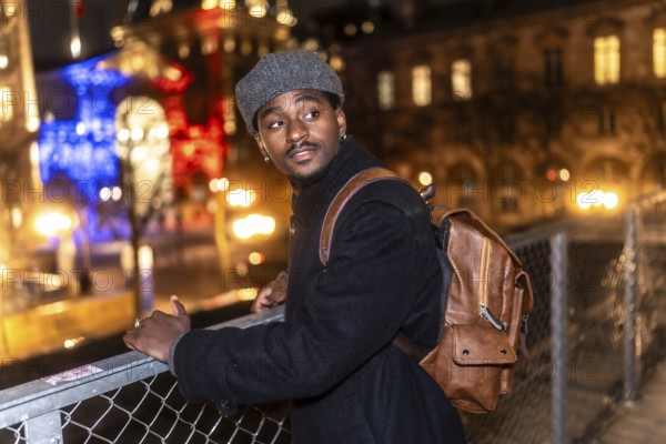 Young black man wearing a coat and a beret, carrying a backpack, standing on a bridge overlooking the illuminated city of paris with the french flag colors in the background at night