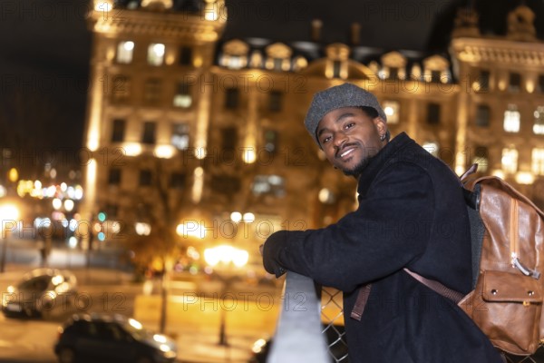 Young man smiling, wearing a beret, coat and backpack, viewing the illuminated city of paris at night from a bridge, with blurry buildings and car lights in the background